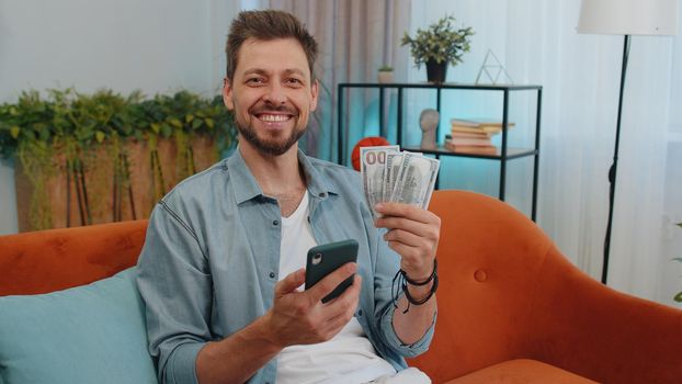 Planning family budget. Smiling happy man counting money dollar cash, use smartphone calculate domestic bills at home. Joyful young guy satisfied of income saves money for planned vacation, gifts