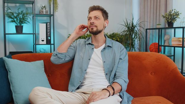 Happy relaxed overjoyed young guy in wireless earphones dancing on couch at home choosing listening favorite energetic disco rock n roll music in smartphone entertaining. People weekend activities
