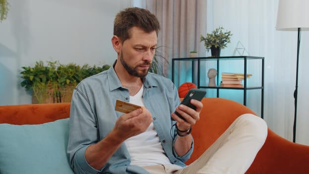 Handsome man using credit bank card and smartphone while transferring money, purchases online shopping, order food delivery at modern home apartment indoors. Young guy in living room sitting on couch