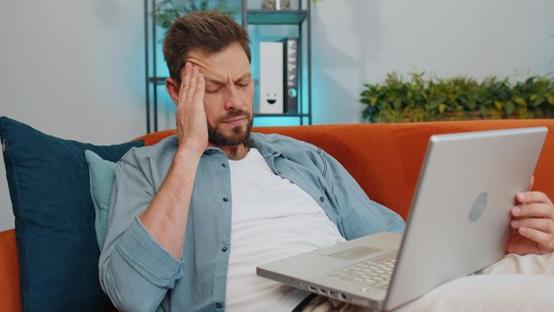 Tired caucasian adult man freelancer use laptop, suffering from headache problem tension and migraine, stress. Young guy works on notebook, sends messages, makes online purchases, watching movies
