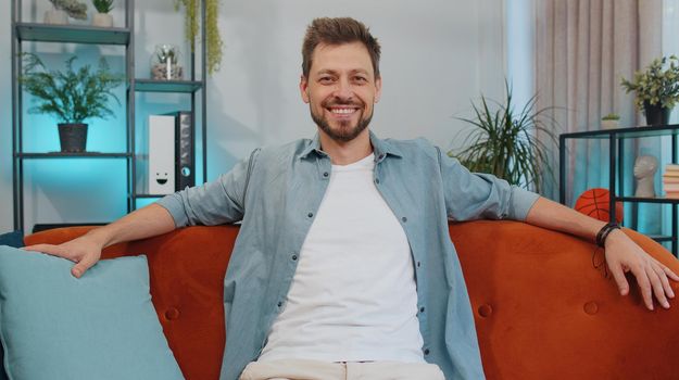 Close-up portrait of happy smiling caucasian adult man in shirt, looking at camera, celebrate good news. Young guy indoors isolated at home in living room sitting on orange couch. Male nature beauty