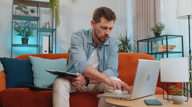 Man accountant working with financial graph documents, calculate taxes for charts and graph reports of home income on couch. Young businessman working on laptop computer, planning expenses budget