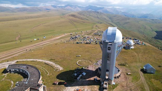 The Assy-Turgen Observatory is high in the mountains. There is a tent camp next to the observatory. Large cumulus clouds in a blue sky. Yellow-green hills, forest in places. Top view from a drone