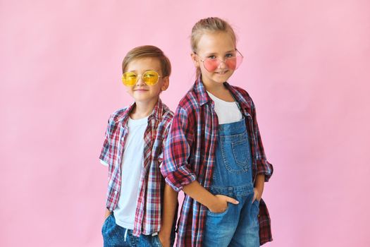 Cute stylish little girl and boy in color sunglasses dressed in plaid shirts, looking at camera and laughing while standing on pink background with copy space.