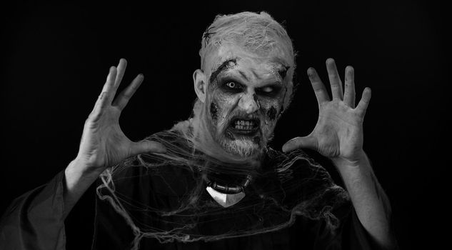 Frightening man with Halloween zombie bloody wounded makeup, trying to scare, face expressions. Horror theme. Sinister undead guy isolated on studio black background. Voodoo rituals. Fashion body art