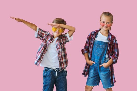 Cute stylish little girl and boy in color sunglasses dressed in plaid shirts, looking at camera and laughing while standing on pink background with copy space.