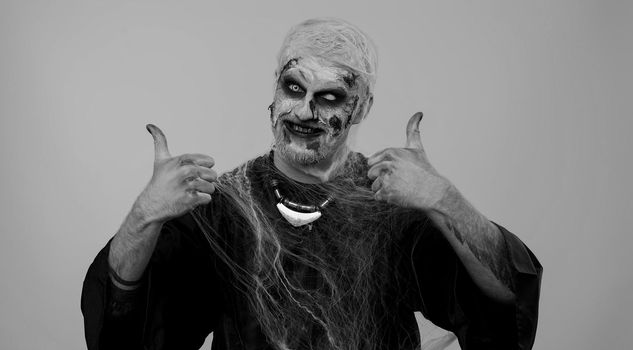 Sinister man with scary Halloween zombie makeup in costume raises thumbs up agrees with something or gives positive reply recommends advertisement likes good. Undead guy with wounded bloody scars face
