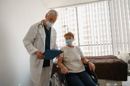 Doctor in mask explaining to a sick patient in wheelchair details of treatment. High quality photo