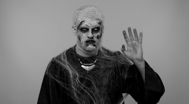 Creepy scary man with bloody scars face, Halloween zombie make-up. Scary wounded undead guy waves hand palm in hello gesture welcomes someone with hospitable expression expresses positive emotions