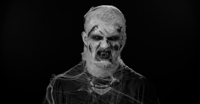 Sinister man with horrible scary Halloween zombie makeup in convulsions making faces, looking ominous, trying to scare. Dead guy with wounded bloody scars face isolated against black wall background