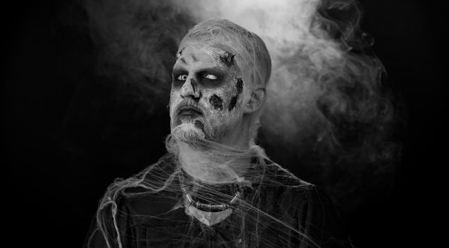 Sinister man in carnival costume of Halloween crazy zombie with bloody wounded scars face screaming, shouting, trying to scare on black room. Horror theme of cosplay wounded undead, beast, monster