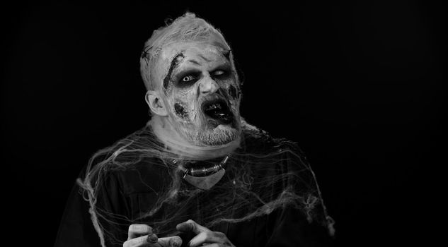 Scary man with horrible scary Halloween zombie makeup in convulsions making faces, looking ominous, trying to scare. Dead guy with wounded bloody scars face isolated against black wall background