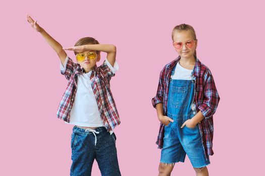 Cute stylish little girl and boy in color sunglasses dressed in plaid shirts, looking at camera and laughing while standing on pink background with copy space.