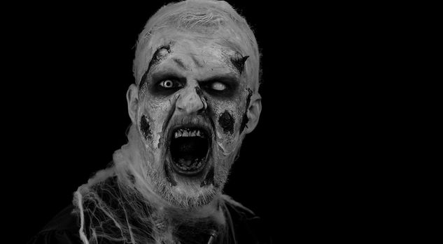 Frightening man with Halloween zombie bloody wounded makeup, trying to scare, face expressions. Horror theme. Sinister undead guy isolated on studio black background. Voodoo rituals. Fashion body art
