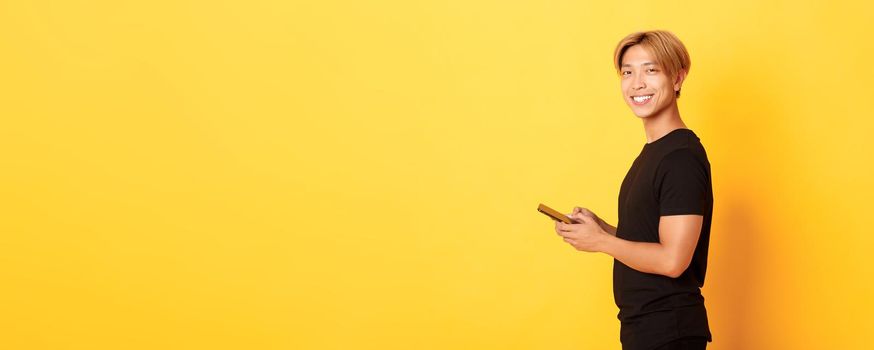 Portrait of handsome stylish asian guy in black outfit, using mobile phone and turning head at camera with satisfied smile, yellow background.