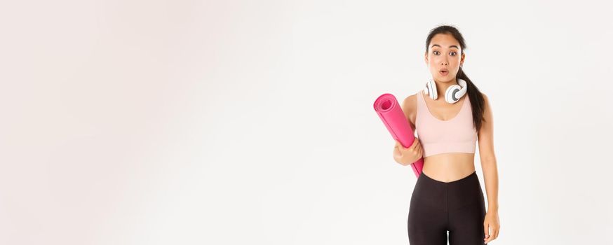 Sport, wellbeing and active lifestyle concept. Confused and shocked asian fitness girl, cant understand why gym closed, holding rubber mat and looking surprised at camera, white background.