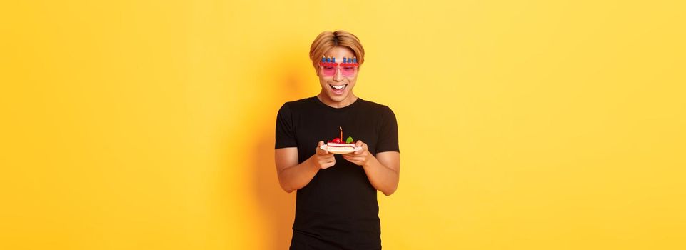 Excited handsome asian guy in party glasses, looking hopeful at birthday cake as celebrating b-day, making wish on lit candle, standing yellow background.