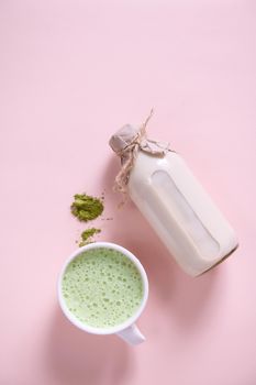 Flat lay. Vertical studio shot of a white ceramic cup of whipped matcha latte with foam, heap of powdered green tea, bottle of healthy organic wholesome raw vegan milk over pink background. Copy space