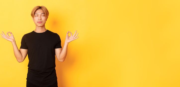 Portrait of relaxed and peaceful handsome asian man with blond hair, close eyes and meditating, attend yoga, standing yellow background.