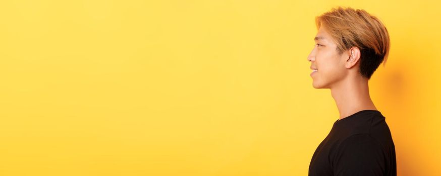 Profile of stylish handsome asian guy with fair hair looking left and smiling, standing over yellow background.