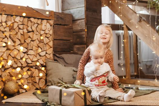Little brother and sister play on Christmas eve in a beautiful house decorated for the New Year holidays. Children are playing with a Christmas gift. Scandinavian-style interior with live fir trees and a wooden staircase.