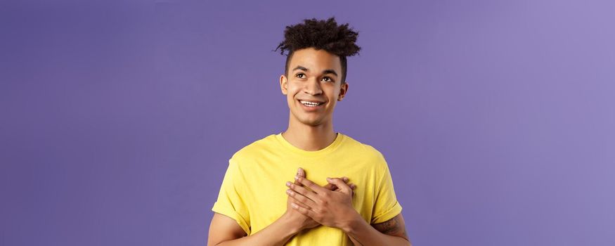 Close-up portrait of touched, romantic attractive man, hold hands on heart, remember nice lovely memory, looking up daydreaming, smiling pleased, appreciate something, purple background.