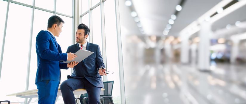 Two business people talk project strategy at office in widen view meeting room. Businessman discuss project planning with colleague at modern workplace .