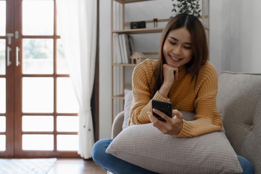 Cheerful asian young woman using mobile phone while sitting on a sofa at home.