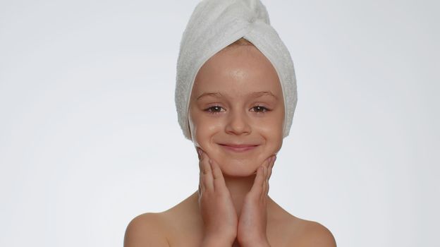 Lovely young child girl after bath in towel on head applying cleansing moisturizing cream. Teenager kid face skincare healthy treatment, natural creme cosmetics. Female portrait. Perfect fresh clean