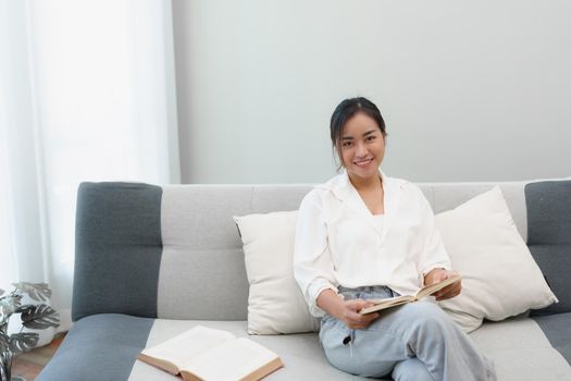 Portrait of a young Asian woman reading a book on the sofa to relax.