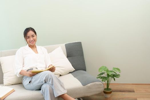 Portrait of a young Asian woman reading a book on the sofa to relax.