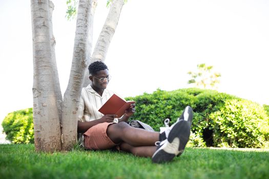 African american university student relaxing on campus.Young black man reading book outdoors. Literature and leisure time concepts. Copy space.