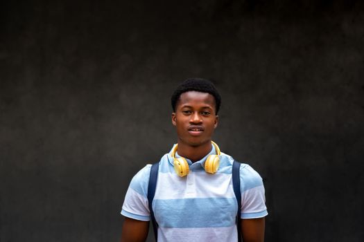 Portrait of teen African American student boy looking at camera outdoors on dark concrete wall background. Copy space. Education concept.