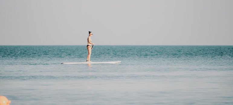 Silhouette of woman standing, surfing on SUP board, confident paddling through water surface. Idyllic sunset or sunrise. Sports active lifestyle at sea or river.