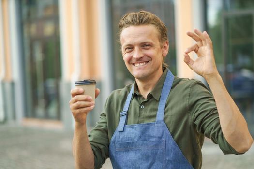 Handsome barista man, guy holding paper cup with fresh made coffee gesturing OK sign with other hand and smile standing outdoors with windows shop on background. Freelancer barista in apron.