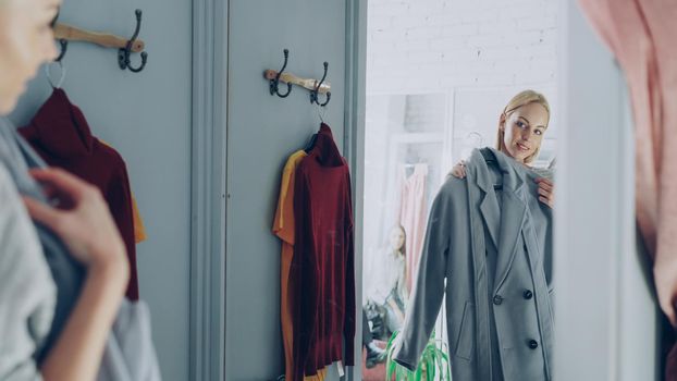 Attractive young woman is trying elegant coat while standing in fitting room is clothing boutique. She is looking at herself in large mirror, slightly turning and smiling.