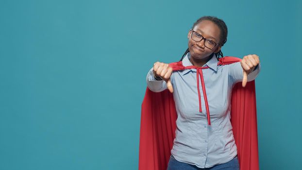 Portrait of woman giving thumbs down and wearing red cape to be cartoon character superhero with comic costume. Doing dislike and disapproval gesture with powerful look, action cloak.