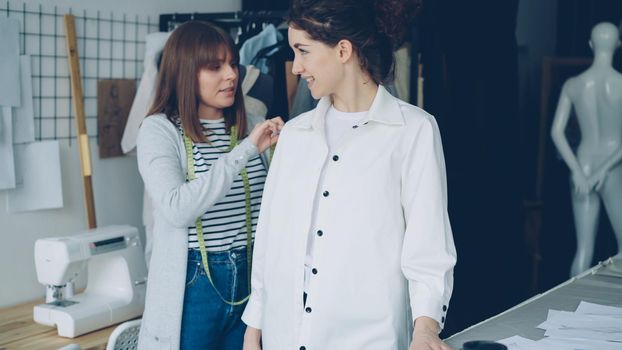 Pretty customer is trying on tailored shirt and sharing her opinion while female clothing designer is measuring and checking new garment and taking to client in light studio.