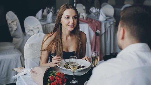 Cheerful attractive girl in beautiful dress is talking to her boyfriend and laughing while having dinner together in restaurant. Conversation, eating out and happy people concept.