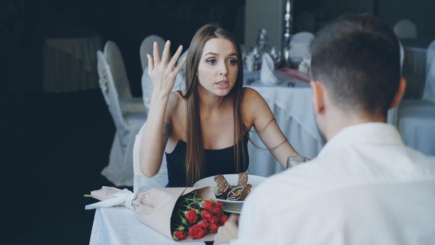 Angry young woman is quarreling with her lover while dining in restaurant, shouting and gesturing emotionally. Bouquet of flowers and plate with food are visible.