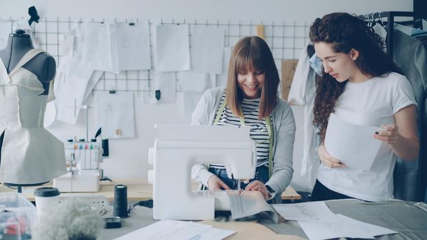 Young woman is working with sewing machine and checking stitches when her colleague is coming to her with sketch. Women are looking at fabric and talking. Teamwork in fashion industry concept.