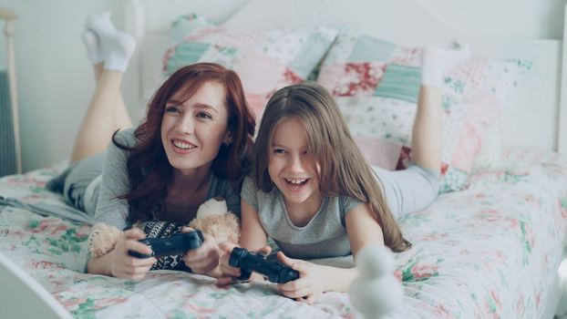 Attractive happy mother with little daughter laughing and have fun while playing computer console games on TV lying on bed at home in morning