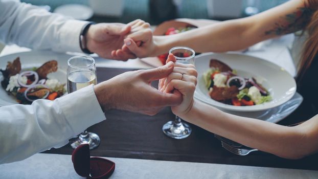 Close-up shot of young lovers touching and holding hands at romantic dinner in classy restaurant. Table with sparking champagne glasses, flatware and food in background.