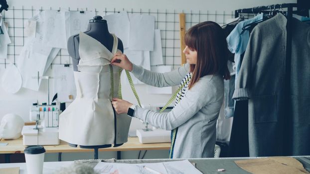 Young seamstress is measuring clothing pinned to tailoring dummy with measure-tape. Attractive woman is concentrated and thoughtful. Studio is light, modern, with many sewing tools and items.