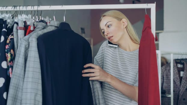 Close-up shot of beautiful young lady talking on mobile phone and choosing clothes in women's clothing store. She is examining colourful stylish garments thoroughly, checking price and smiling.