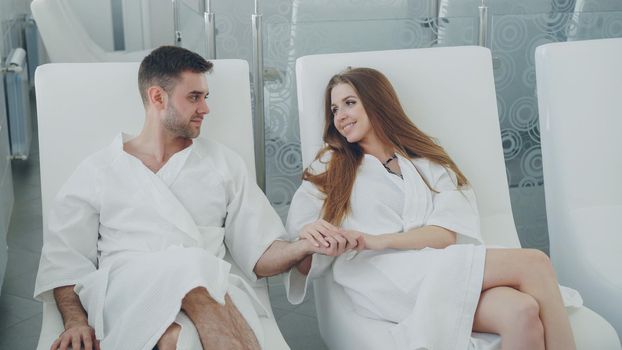 Handsome bearded man and his pretty girlfriend wearing bathrobes are sitting in chairs in relaxation room in day spa, drinking cocktails and talking. Relationship and wellness concept.