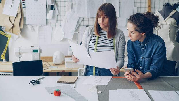 Attractive young women working in design industry are comparing sketches and talking about them while working in modern tailor shop. Productive teamwork concept.