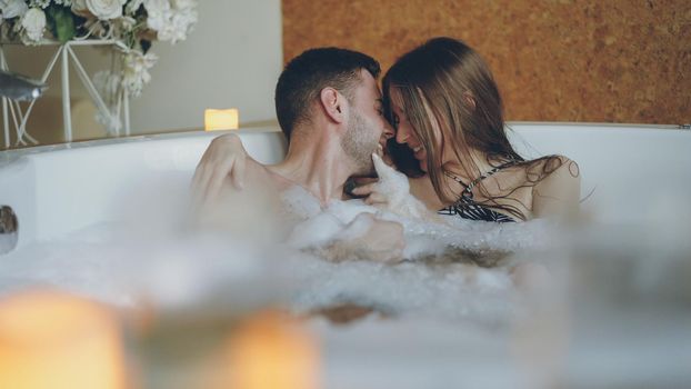 Adorable cheerful young couple is having fun in bubbling hot tub playing with foam, talking and kissing. Romantic relationships, happy people and wellness concept.