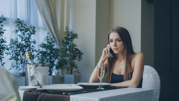 Angry beautiful woman is calling her boyfriend on mobile phone while waiting for him sitting alone in restaurant. Girl is impatient, nervous and offended.