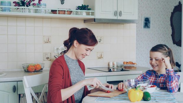 Happy young mother cooking vegetables cutting tomatoes and talking to her cheerful daughter in the kitchen at home. Family, cook, and people concept